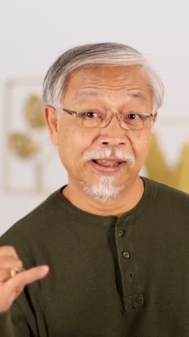 Smiling senior Asian man with glasses makes playful phone call gesture in bright indoor setting.