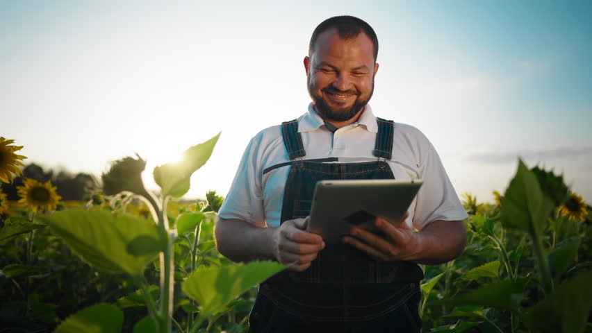 Portrait of cheerful farmer with tablet in beautiful sunflower field in summer. Modern agricultural technology and innovation in agribusiness, inspired agronomist admiring rural scenery in farmland
