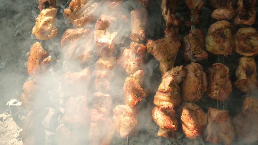 Top view of appetizing juicy shashlik pieces on multiple skewers grilling over hot coals. The cook rotates skewers for even roasting. Traditional Armenian khorovats barbecue cooking outdoors.