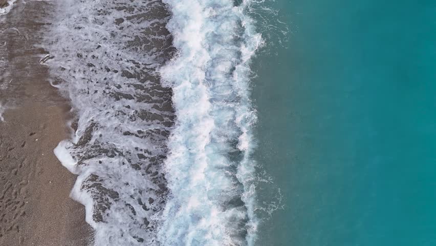 A mesmerizing 4K top-down aerial shot of a gentle white wave rolling up a pristine pebble beach creating a beautiful and calming natural gradient from turquoise sea to shore
