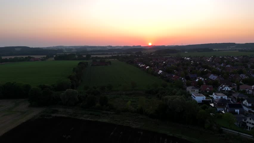 Drone flight along agricultural fields in rural area near luxury houses at golden sunset. Wide shot. Houses and homes in suburb. Cropland and farmland in serene atmosphere. Summer season.