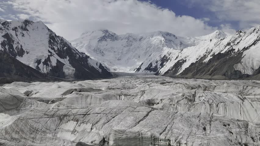 Stunning aerial drone footage of a massive glacier with the majestic Peak Pobeda (Victory Peak) towering in the background. High-altitude mountain landscape with snow-covered peaks, rugged terrain
