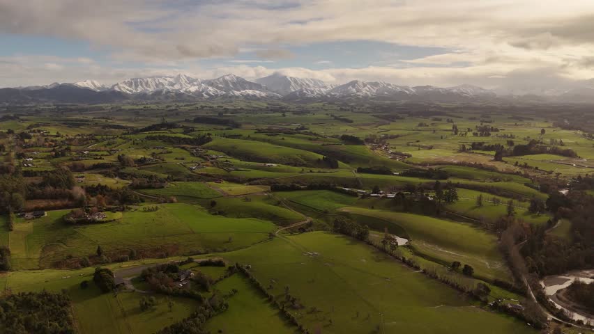 Lush green rolling hills and farmland, majestic snow-capped in background under cloudy sky, Canterbury, New Zealand. Aerial