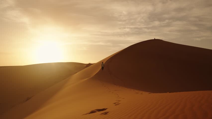 A solitary traveler ascends a golden sand dune in the Sahara Desert during sunrise, leaving a winding trail of footprints under the warm Moroccan sky.