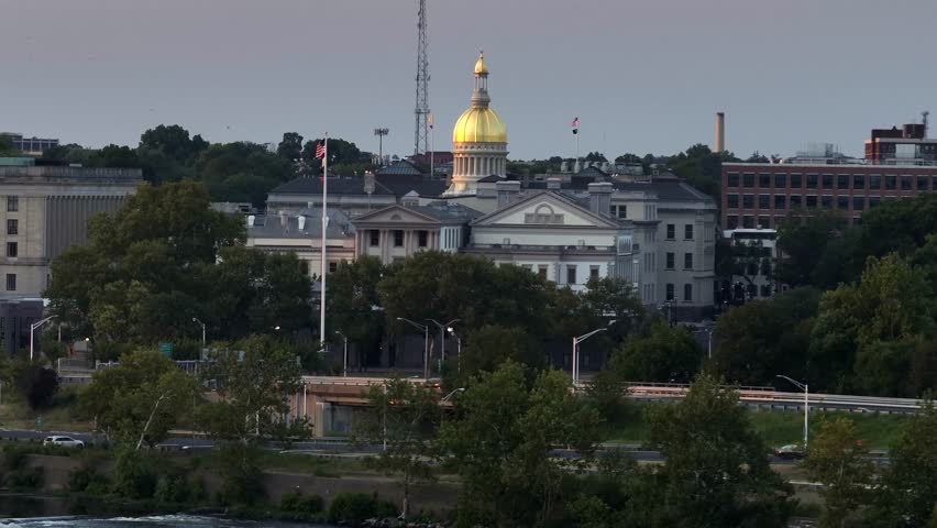 New York State capitol building with golden cupola in Trenton city. Cars on road along river. Waving American flag. Aerial rising wide shot.