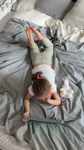 Little girl lying on her parents bed and watching cartoons on a tablet. Concept of screen time, digital entertainment and relaxation during early childhood. High quality 4k footage
