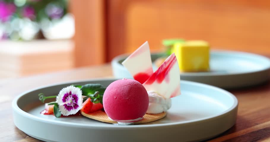 Gourmet dessert plate featuring vibrant raspberry mousse, artistic chocolate shards, fresh fruits, garnished with edible flowers on wooden table. Elegant culinary presentation in natural setting.