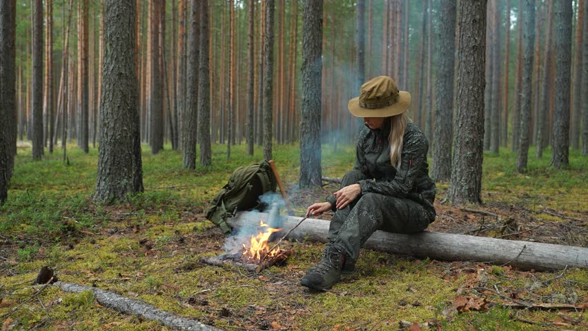 A young woman in hiking clothes warms herself by a campfire in the forest. A female hiker sits beside a woodland campfire. Relaxing in the woods by a campfire. The concept of bushcraft and outdoor