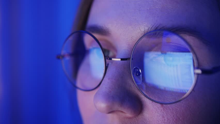 Close up of concentrated woman wearing glasses looking at laptop screen while working. Reflection of laptop monitor in glasses. 