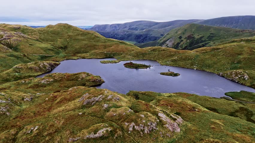Aerial Mid angle wide angle shot of angle tarn lake atop mountain in Lake District England with mountains and valleys in background in summer with clouded sky