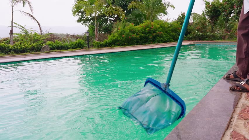 Poolside worker sweeps floating dirt with a cleaning pole, Man uses long net to remove debris from swimming pool, Caretaker moves mesh across sparkling water, Guy on edge maintain aquatic hygiene