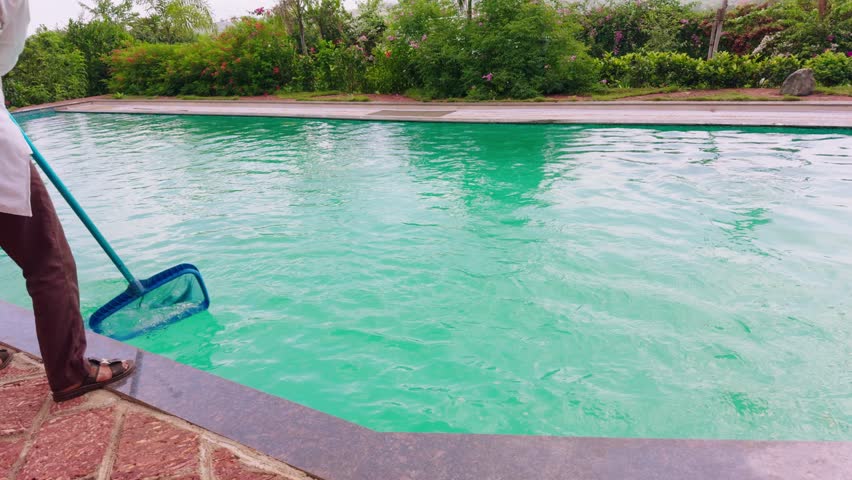 Man uses long net to remove debris from swimming pool, Poolside worker sweeps floating dirt with a cleaning pole, Caretaker moves mesh across sparkling water, Guy on edge maintain aquatic hygiene