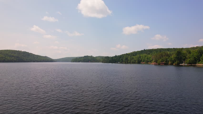 Low drone view of Baptist Lake surrounded by green forest and vacation cottages, blue sky with scattered clouds in Canada. Clean fresh waters of Northern Ontario near Bancroft.