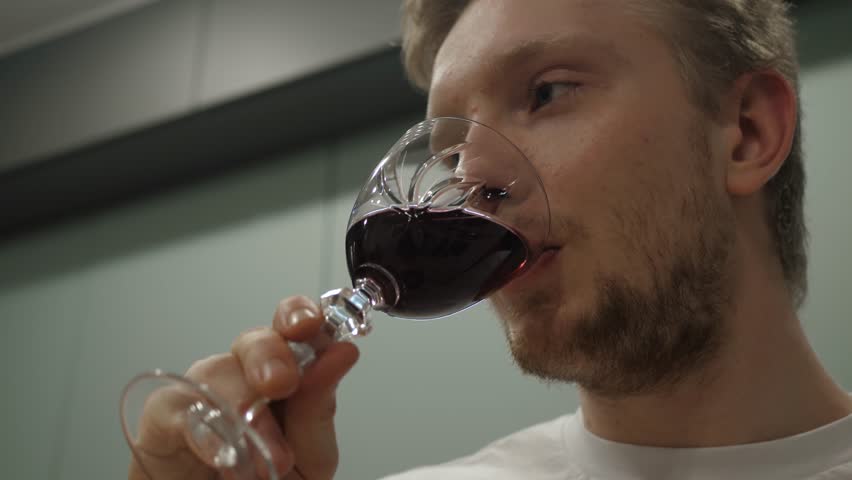 Thoughtful young man enjoying generous glass of red wine in modern kitchen illustrating moment of relaxation and pleasure, close up
