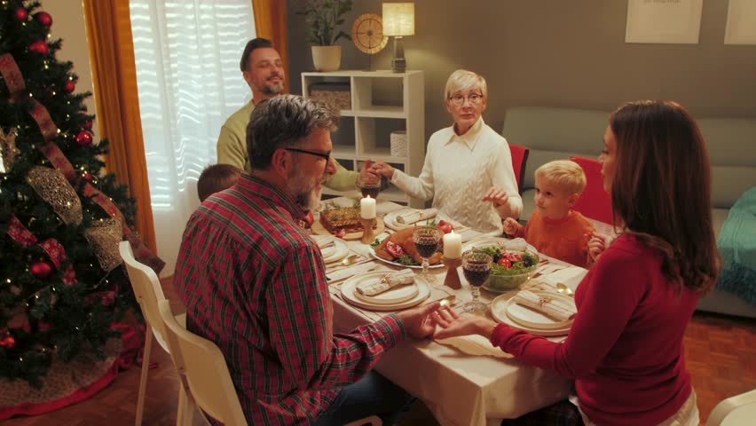 Multigenerational caucasian family gathering around dining table, joining hands in prayer before sharing festive christmas meal, expressing spiritual connection and familial love