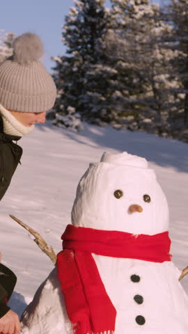 SLOW MOTION, CLOSE UP, PORTRAIT: Happy woman smiles after putting a black hat on a funny snowman she built in an empty snowy meadow. Caucasian female is having fun making a snowman on sunny winter day