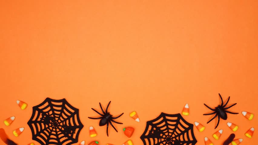 Halloween bottom border of scattered candy and spider webs. Top view stop motion loop video over an orange background with copy space.