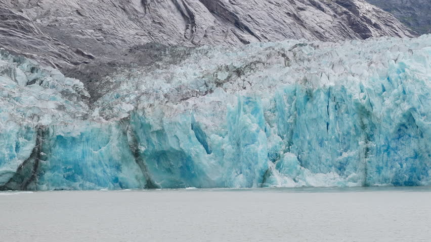 This is a close-up shot of a whole blue ice wall calving off the Dawes Glacier in Endicott Arm near Juneau Alaska. It shows white seagulls flying around showing scale and has a huge splash and wave.