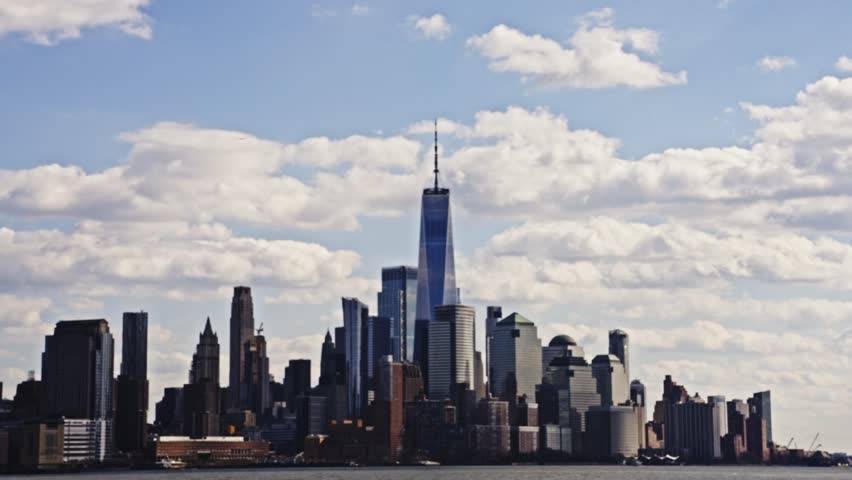 Dramatic view of One World Trade Center rising above the Lower Manhattan skyline on a partly cloudy day, seen across the Hudson River.