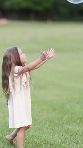 Close-up of a child in white dress watching a large soap bubble in the air on a grassy field. 