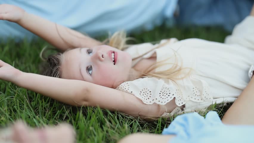 A cute young girl in a white dress lying on her back on green grass during summer.