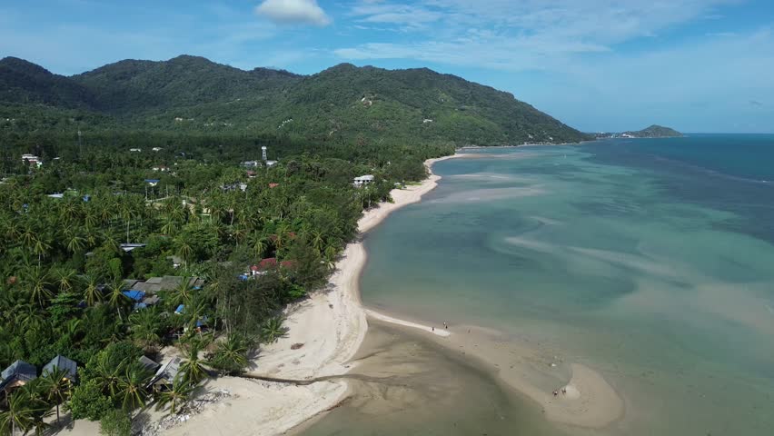 Aerial drone footage of Baan Tai Beach on Koh Phangan, Surat Thani, Thailand, showing palm trees, tropical sandbanks, turquoise sea, green hills, and people swimming and relaxing in the water.