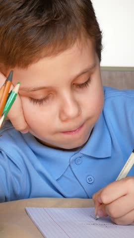 A cute boy of 6-7 years old writes with a pen in a notebook, resting his head. Next to it is a glass with colored pencils. Distance learning during quarantine