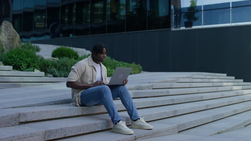 Happy young student celebrates while using a laptop outside, sitting on modern city steps surrounded by greenery and buildings.