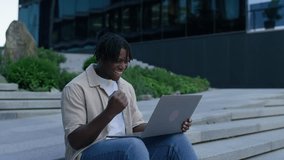 Happy young student celebrates while using a laptop outside, sitting on modern city steps surrounded by greenery and buildings. - Powered by Shutterstock - Get 15% off with code: PIKWIZARD15