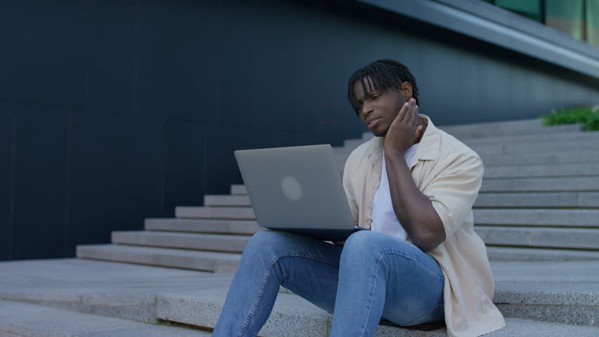 Young student sits outdoors on steps with a laptop, looking puzzled and stressed while struggling with work or studies in the city.