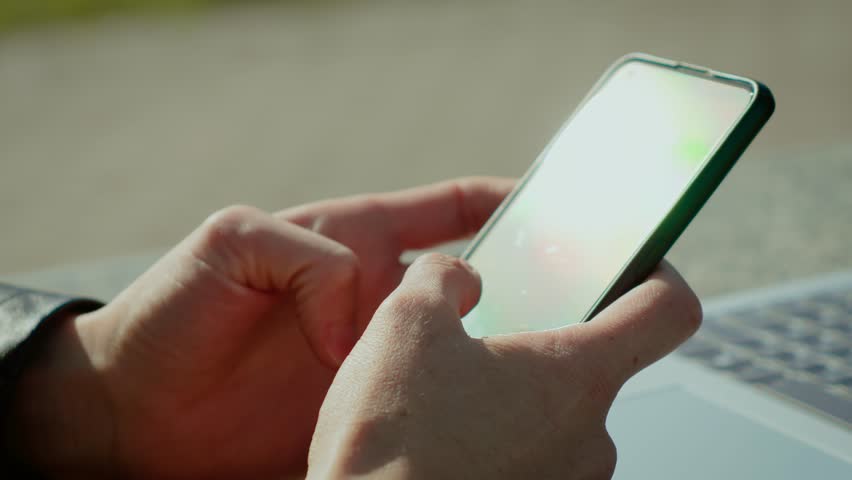 Woman hands holding smartphone typing next to laptop with sun reflections in screen. Fingers using mobile phone beside computer while sunlight glaring. Person interacting with cellphone near notebook