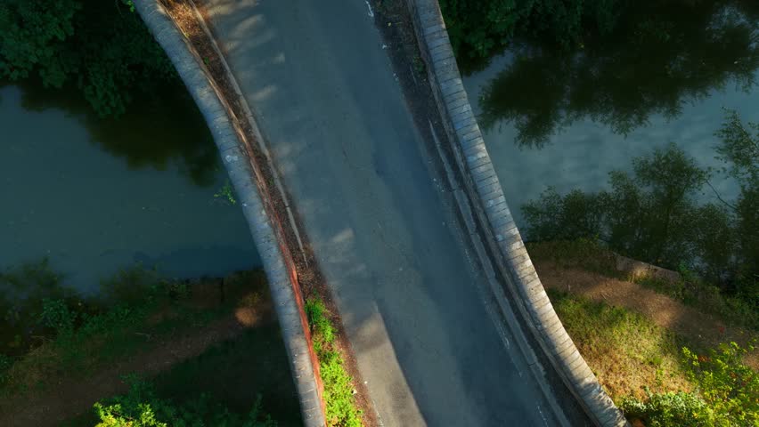 Old stone bridge crossing tranquil forest river. Narrow passage over calm water in lush greenery top down view. Rustic road leading through woodland over stream