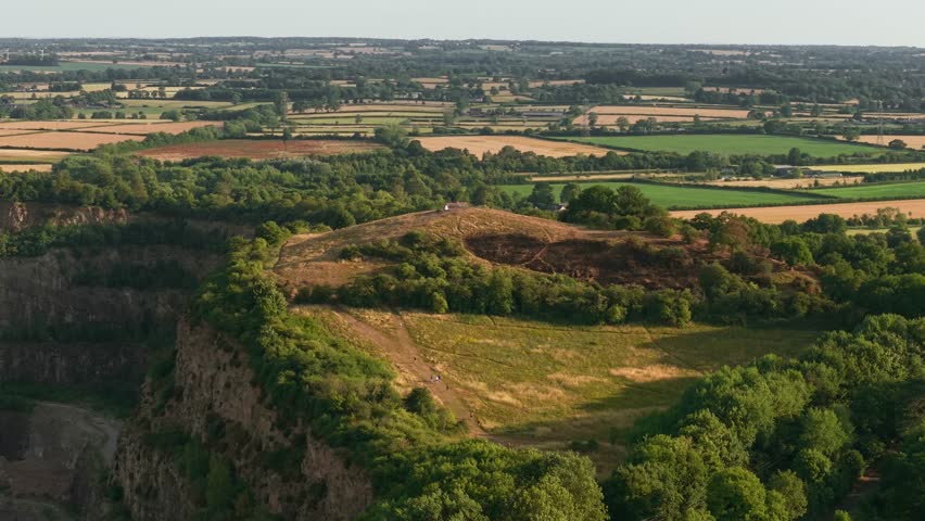 Hilltop standing above quarry rim surrounded by rural landscape. Memorial mound rising over excavation edge bordered by farmlands. Elevated landmark overlooking mining site framed by fields and trees