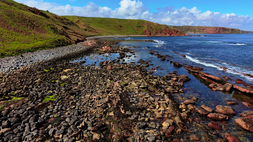 Waves crashing over scattered rocks on North Sea shore in Scotland. Swells breaking along jagged red formations beneath rolling hills. Foam swirling through shallow water near pebbled coastline