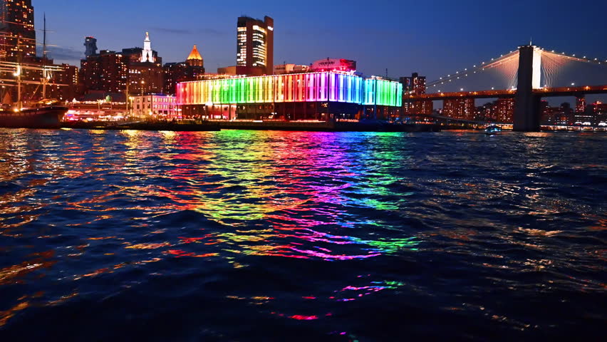 Beautiful rainbow illumination of Pier 17 Bar reflecting in the dark riverscape. River cruise approaching the Brooklyn Bridge at night. New York, USA.