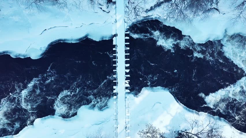 Aerial drone view of fast river with suspension foot bridge. Snow winter forest in Finland 