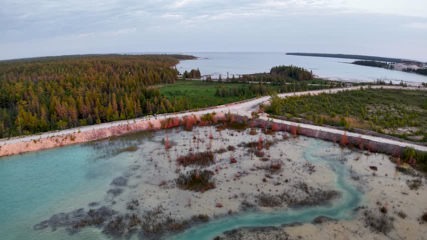 Aerial drone view of a breakwater wall stretching into turquoise coastal waters with a forested shoreline in the distance.