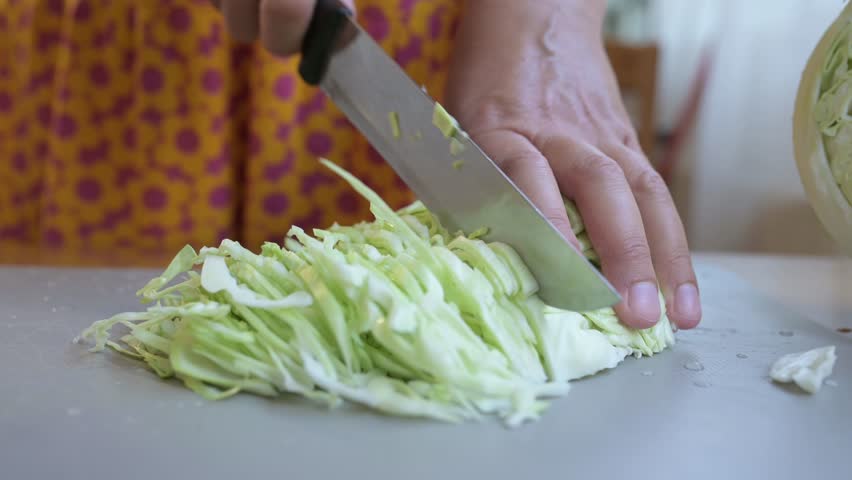 Woman cutting fresh cabbage on a kitchen table. Close-up of hands slicing the vegetable on a cutting board, preparing ingredients for cooking or salad at home