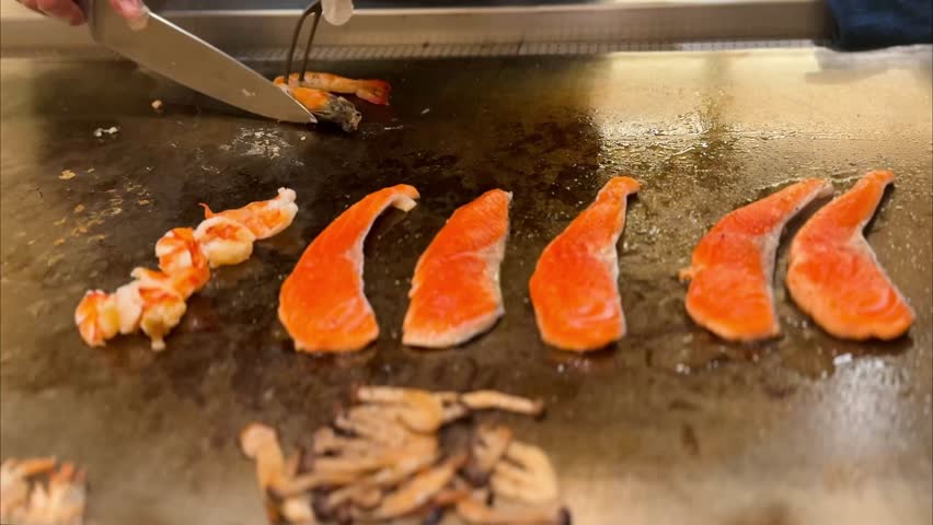 
Close-up of salmon fillets grilling with shrimp being cut on a hibachi teppanyaki griddle beside mushrooms. Authentic Japanese cooking action, perfect for food, cuisine, and restaurant themes.