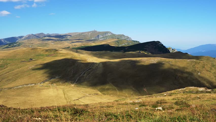 A timelapse of a large mountain range with rolling hills and clouds casting long shadows under a clear sky.