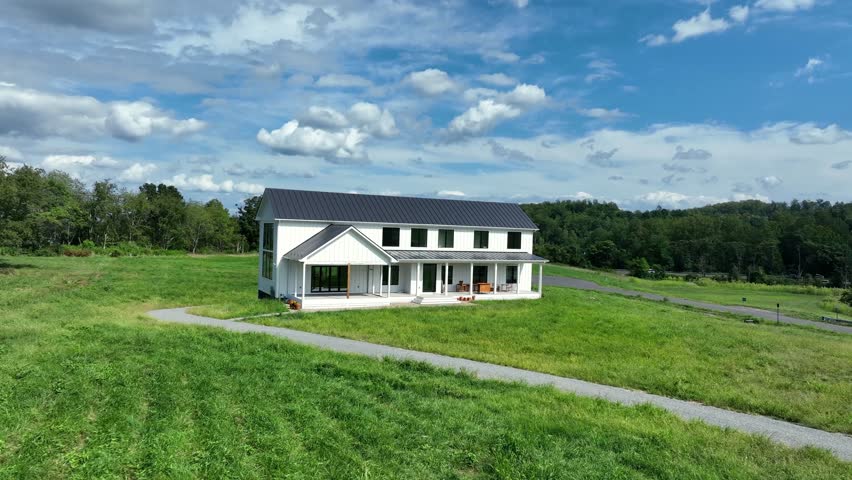 Modern white farmhouse with large front porch, surrounded by green fields and forest, under a bright blue sky with clouds. rural American countryside architecture in Virginia state. Summer day. Aerial