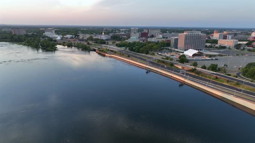 Trenton cityscape near River during golden sunrise. Aerial wide shot. Traffic on shoreline road. Apartment blocks and offices in American city downtown.