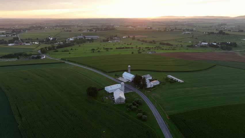 Rural intersection leading to Americn farmstead with rustic silo storages during golden sunrise. Quiet and quaint suburb district of town in Pennsylvania. Aerial approaching wide shot. Green cropland