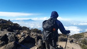 Solo hiker walking with trekking pole on rocky mountain ridge above sea of clouds under bright blue sky, showcasing extreme nature adventure and solitude - Powered by Shutterstock - Get 15% off with code: PIKWIZARD15