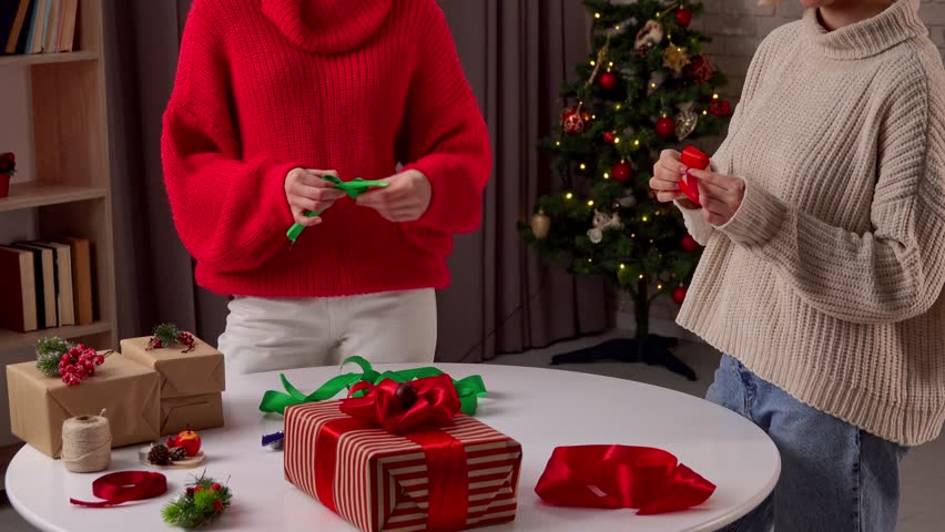 Two girlfriends joyfully participate in holiday preparations, wrapping homemade gifts with colorful ribbons in a cozy living room adorned for Christmas. The atmosphere is warm and festive.
