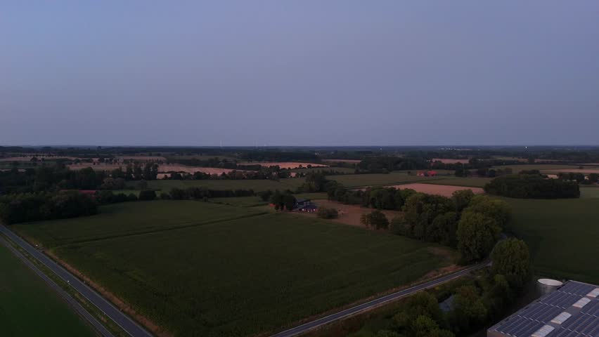 Dusk scene in American countryside. Farmstead with stable and solar panels on roof. Forest trees and agricultural farm fields after sunset time. Peaceful and quaint landscape. Drone wide shot.