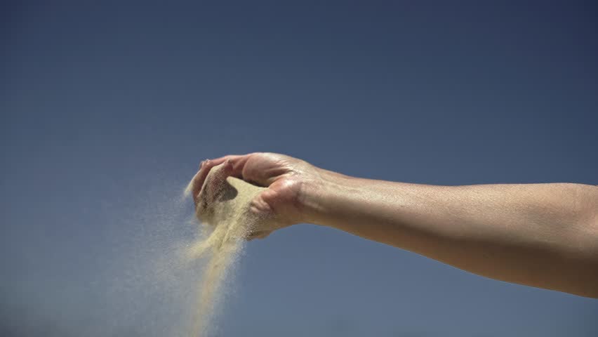 Slow-motion close-up of a woman's hand with sand slipping through fingers against a sky background. Symbolizes fleeting time and life's transience.