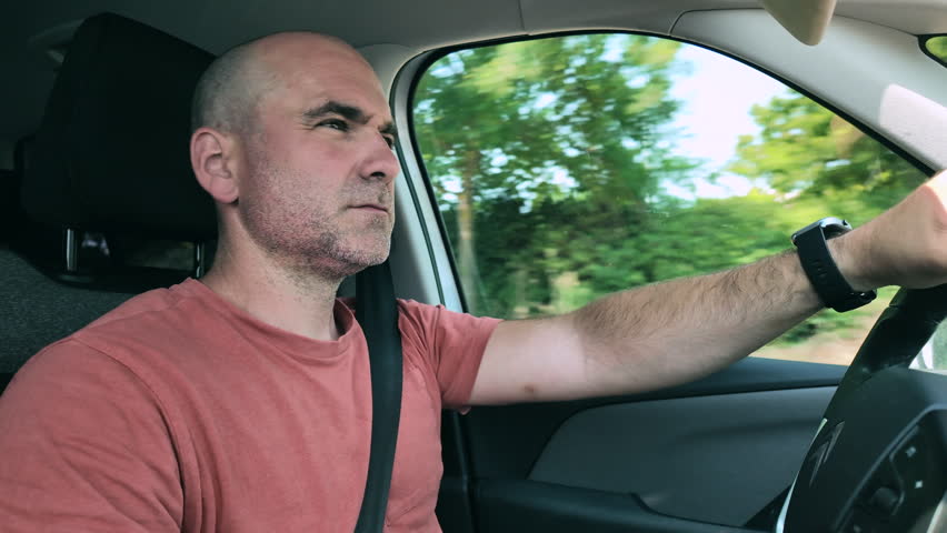 A casually dressed man driving a car, medium profile shot from the inside of the car.