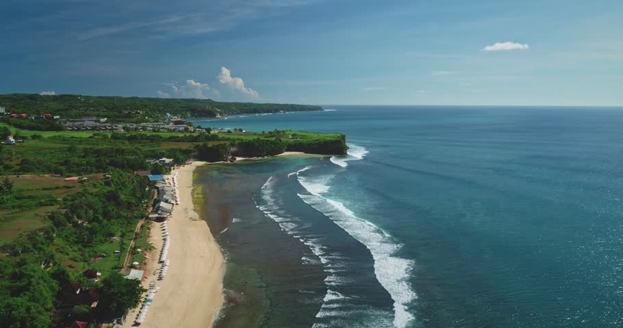 Waves crashing on the shore of Dreamland Beach in Bali, Indonesia, on a sunny day, with beach umbrellas and some vegetation visible from an aerial point of view