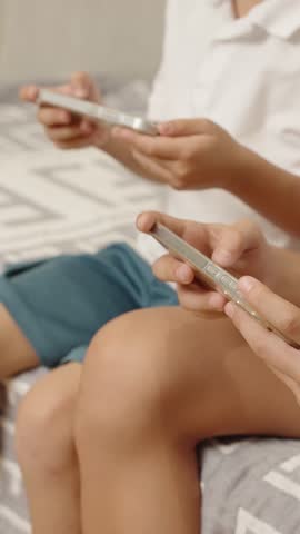 Unrecognized children sitting on bed holding mobile device and engaged in playing games. Two boys tapping screens of smartphones while playing online. Kids entertaining with gadgets in the internet.
